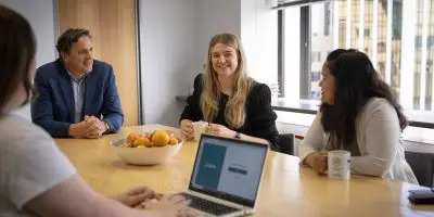 Four individuals collaborate around a laptop, sharing a light moment at a large dining table. A bowl of oranges is placed in the centre of the table
