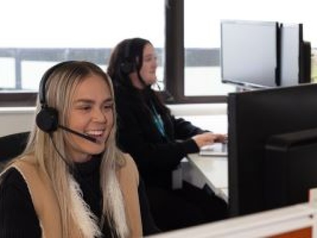 Two women at their desks, wearing headphones and engaged in phone conversations while also interacting with their desktop computers. They exude a sense of happiness and contentment