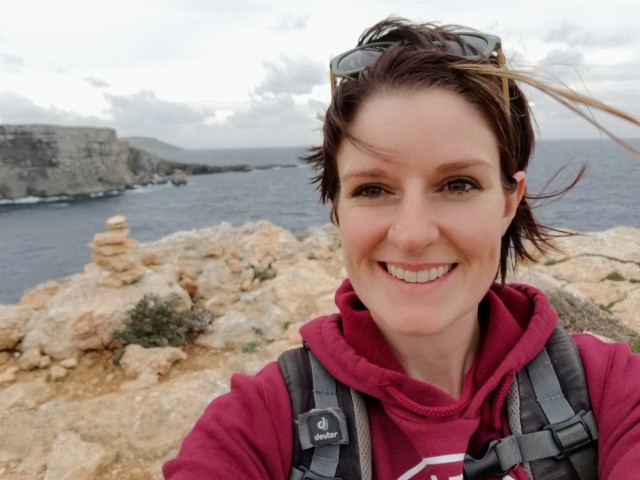 A woman smiling into the camera, with sea and grey skies in the background.