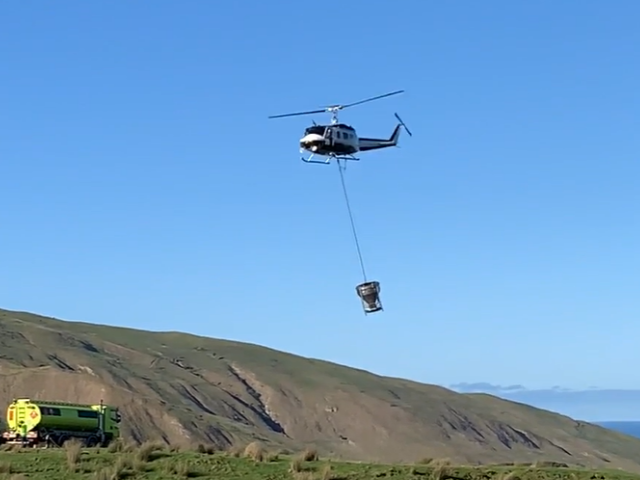 A helicopter flying over hilly terrain, dispensing 1080 bait from a hopper hanging beneath it